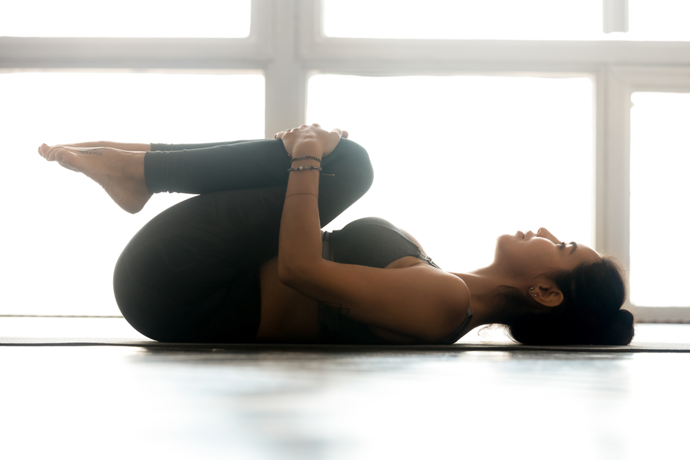 Woman doing knees to chest pose Young sporty attractive woman practicing yoga, doing Knees to Chest exercise, Apanasana pose, working out, wearing sportswear, grey pants, top, indoor full length, at yoga studio