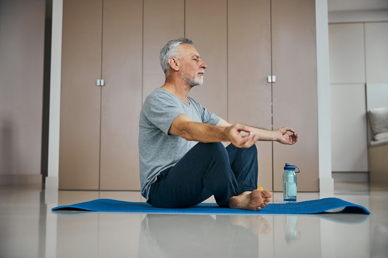 A grey-haired man sitting on a yoga mat with eyes closed, meditating. 
