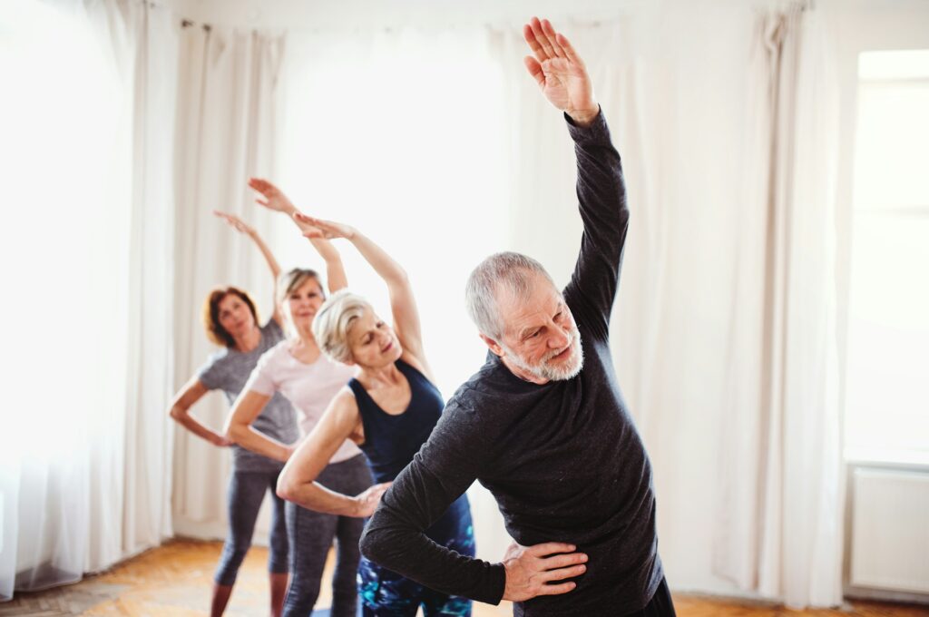 Four people stretching their arms over their heads while standing in a room with white curtains. 